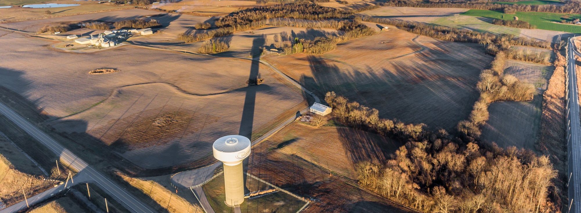 Aerial view of a rural landscape with open fields, scattered buildings, and a tall water tower casting a long shadow across the land. Sparse trees border the fields, and country roads intersect near the bottom left.