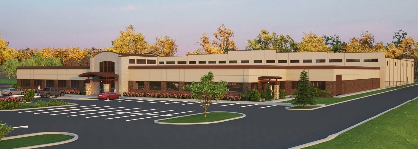 A modern, single-story building with large windows and stone accents is surrounded by trees. There is a parking lot with several cars in front, landscaped with shrubs and small trees. The sky is clear.
