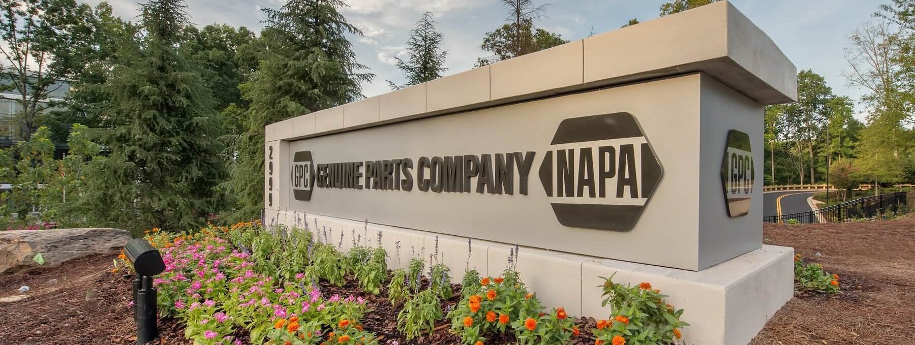 A large outdoor sign reads "Genuine Parts Company (NAPA)" surrounded by colorful flowers, greenery, and trees on a landscaped area.