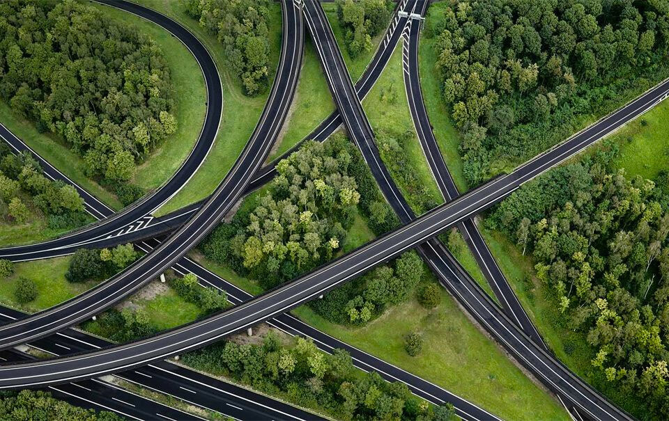 Aerial view of a complex highway interchange with multiple roads and overpasses winding through a dense, green forest. The roads crisscross each other, surrounded by lush trees and grassy areas.