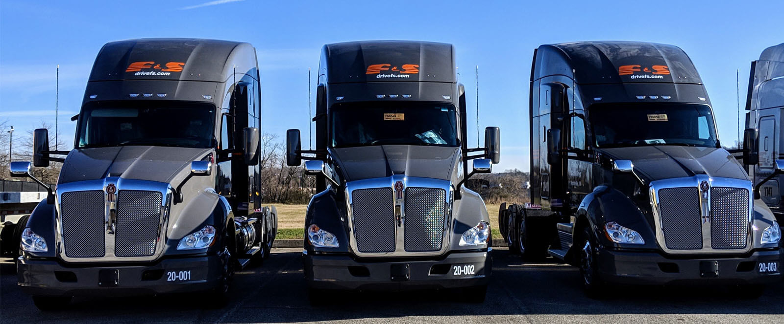Three black ELS semi-trucks parked side by side in a lot under a clear blue sky, facing forward.