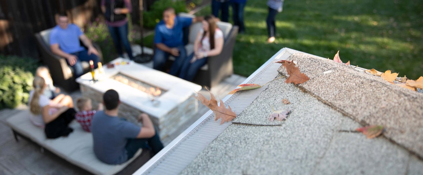 Close-up of a house roof with leaves on a gutter, with a blurred background showing a group of people sitting and talking around a fire pit in a backyard.