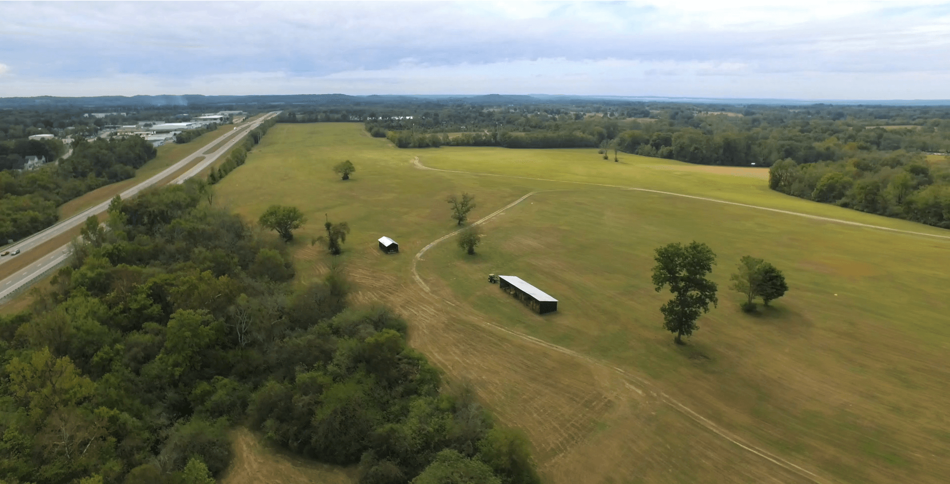 Aerial view of a wide open grassy field with scattered trees, two small rectangular buildings, a curving dirt path, and a nearby highway bordered by forested areas under a mostly cloudy sky.