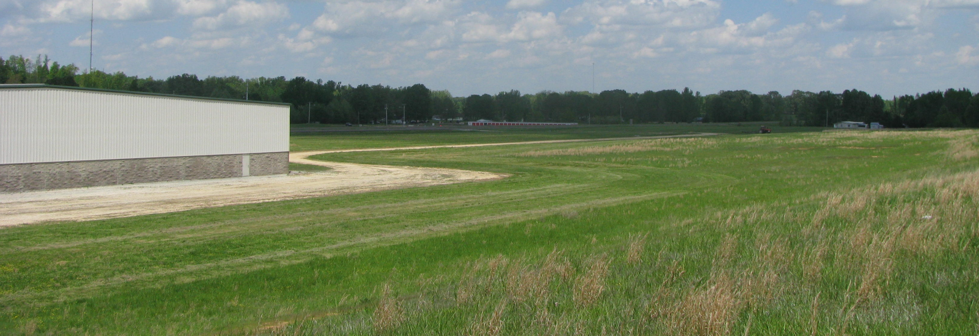 A grassy field with a curved dirt road beside a white building, bordered by trees under a partly cloudy sky.