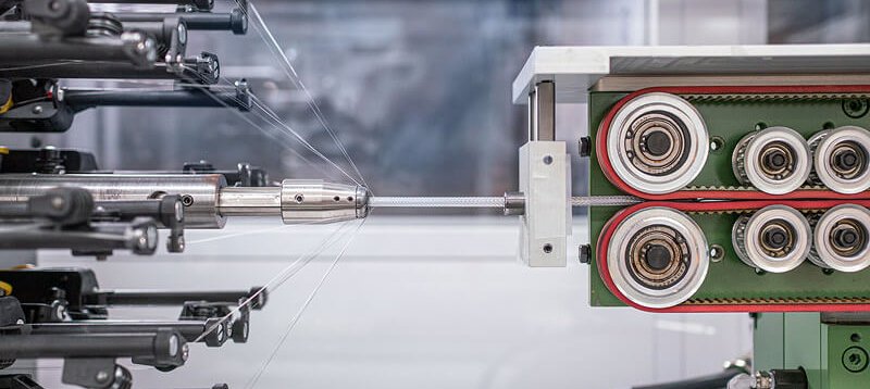 Close-up of an industrial wire braiding machine in operation, showing multiple wires being woven together as they pass through cylindrical guides and rollers inside the mechanical apparatus.