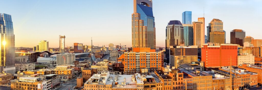 A panoramic view of downtown Nashville, Tennessee at sunset, featuring a mix of modern skyscrapers and historic brick buildings under a clear sky.