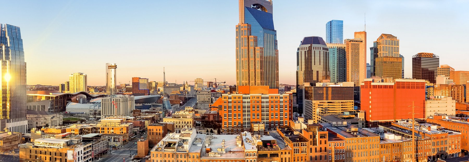 A panoramic view of downtown Nashville, Tennessee at sunset, featuring a mix of modern skyscrapers and historic brick buildings under a clear sky.