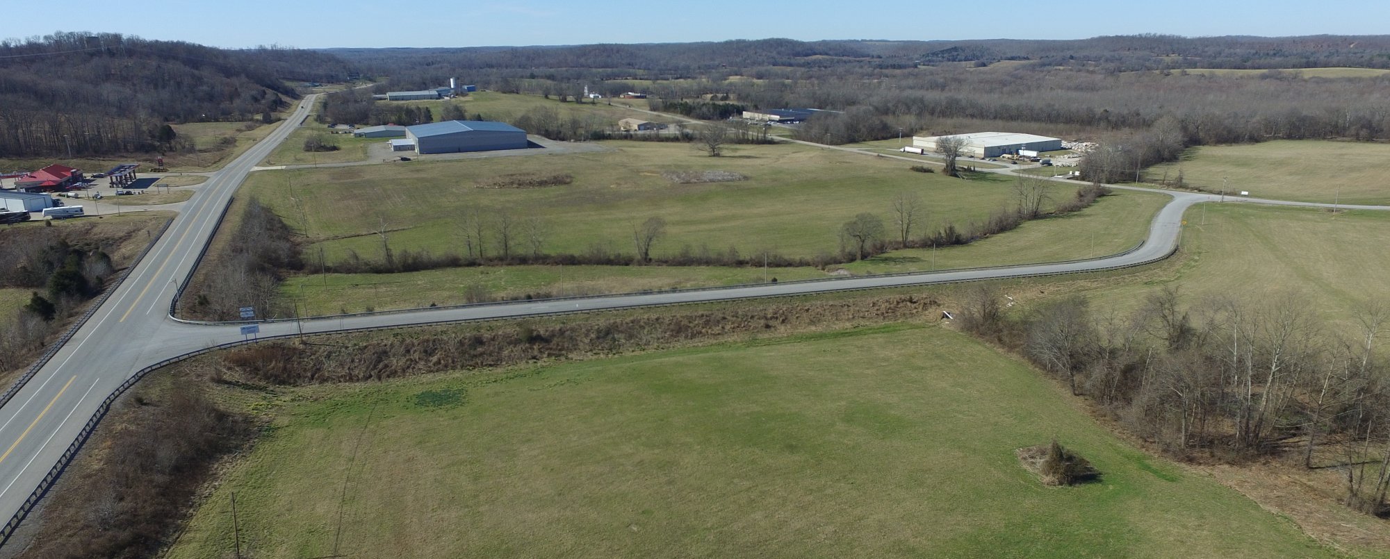 Aerial view of a rural landscape with green fields, a curving road, scattered trees, and several large industrial buildings; forested hills appear in the background under a clear blue sky.