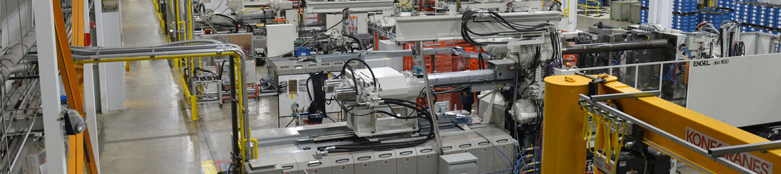 A factory floor with multiple large industrial machines and equipment, including injection molding machines, arranged in rows with visible pipes, wires, and safety barriers.