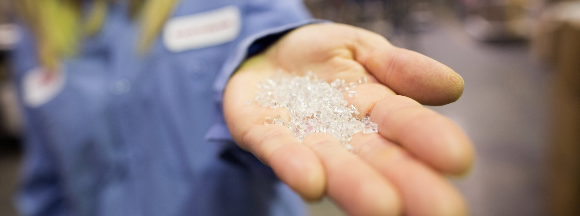 A person in a blue shirt holds clear plastic pellets in their outstretched hand, with the background out of focus.