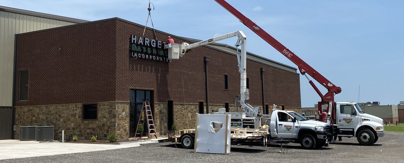 Workers use a bucket truck and crane to install a "Hargett Materials Inc." sign on the exterior of a brown brick and stone commercial building under a clear blue sky. Construction equipment is visible on the ground.