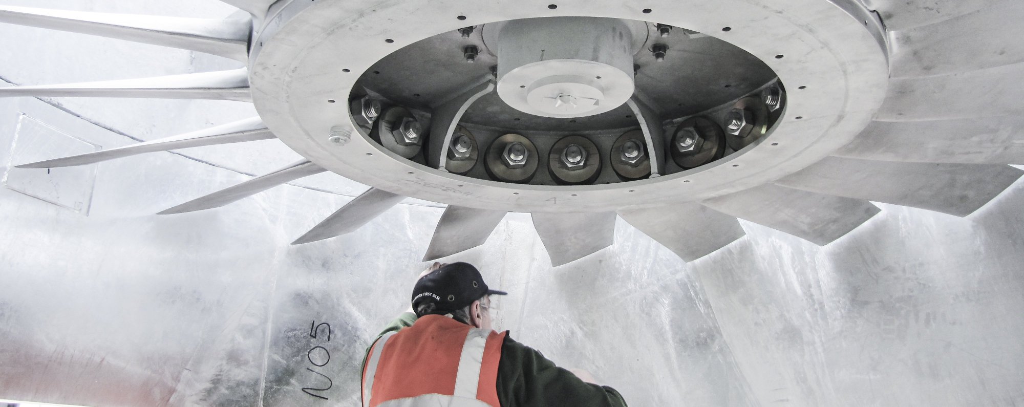 A worker wearing a cap and high-visibility vest inspects the interior of a large, industrial metallic turbine with curved blades and visible mechanical components.