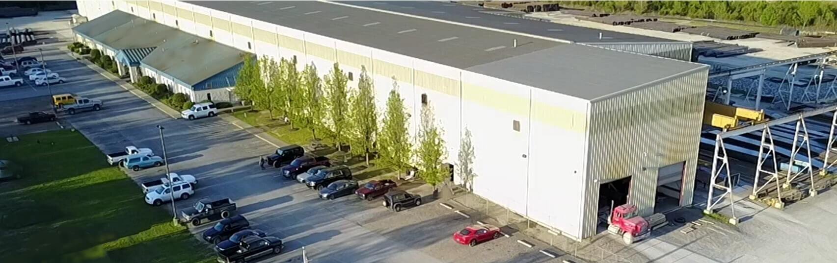 Aerial view of a large industrial warehouse with a long white roof, lined with trees in front, and cars parked in rows along the adjacent parking lot.