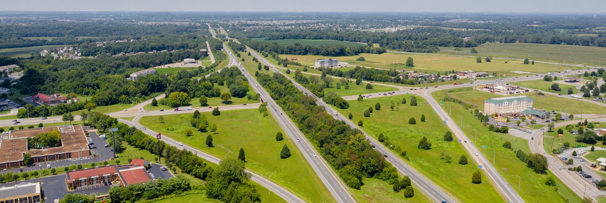 Aerial view of a wide highway with multiple lanes and on- and off-ramps surrounded by green fields, scattered buildings, and trees, stretching into the distance under a clear sky.