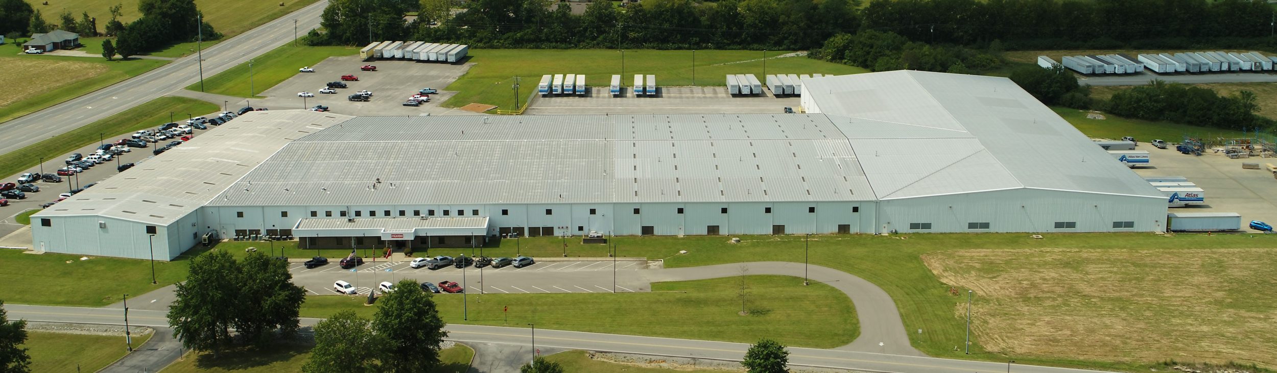 A large industrial warehouse with a light gray roof, surrounded by parking lots with cars and trucks, and open green areas. Multiple loading docks and trailers are visible at the back.