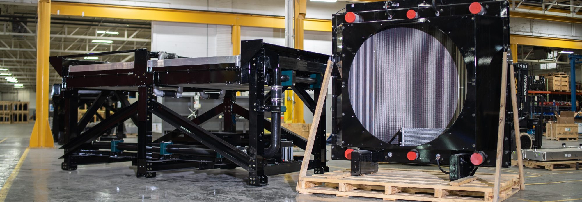 Large industrial metal structures are displayed in a spacious warehouse; one frame stands upright on a wooden pallet, while another lies horizontally in the background, both featuring black steel and metallic mesh components.