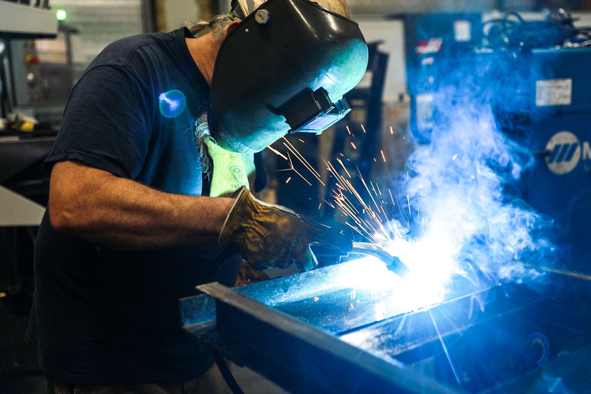 A person wearing protective gear, including gloves and a welding helmet, is welding metal in a workshop. Bright blue sparks and smoke surround the welding area.