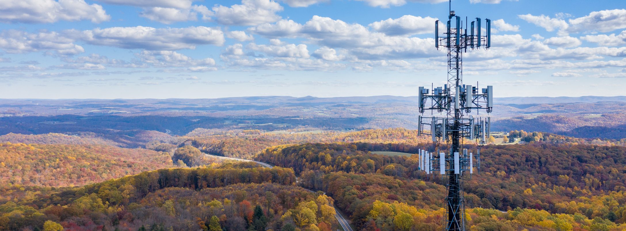 A cell tower stands on a hill above a forest with autumn foliage, under a partly cloudy sky. Rolling hills and a winding road stretch into the distance.