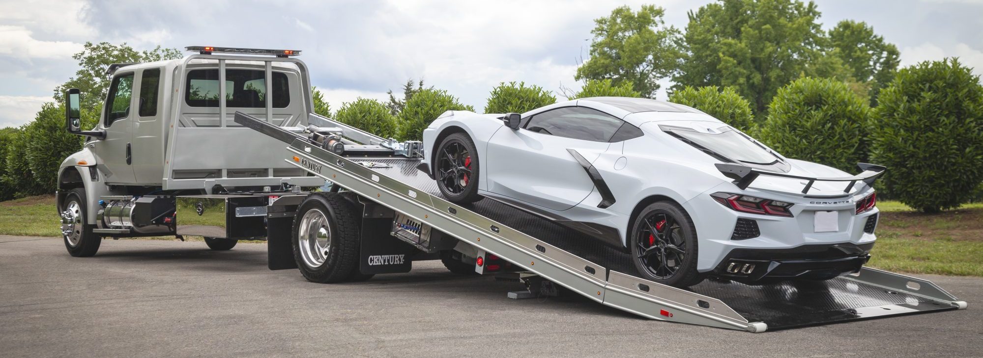 A silver flatbed tow truck with a tilted ramp is loading a white sports car with black accents and red brake calipers. Green trees and cloudy sky are visible in the background.