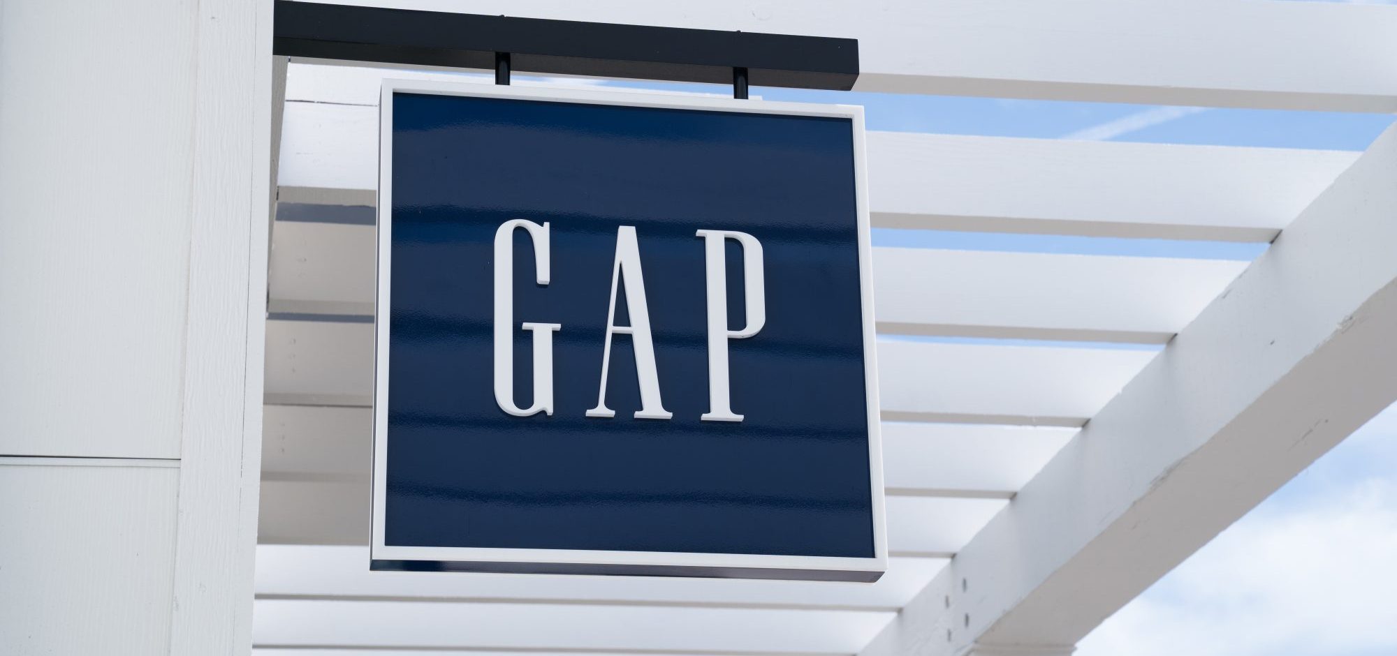 A dark blue square sign with the word "GAP" in large white capital letters hangs from a white pergola structure against a partly cloudy sky.