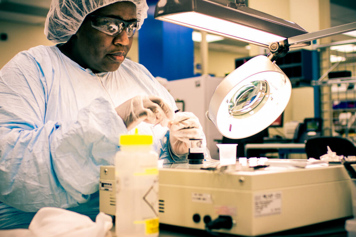 A scientist wearing a hairnet, lab coat, gloves, and safety glasses works with lab equipment under a bright lamp in a laboratory, handling small objects with precision.