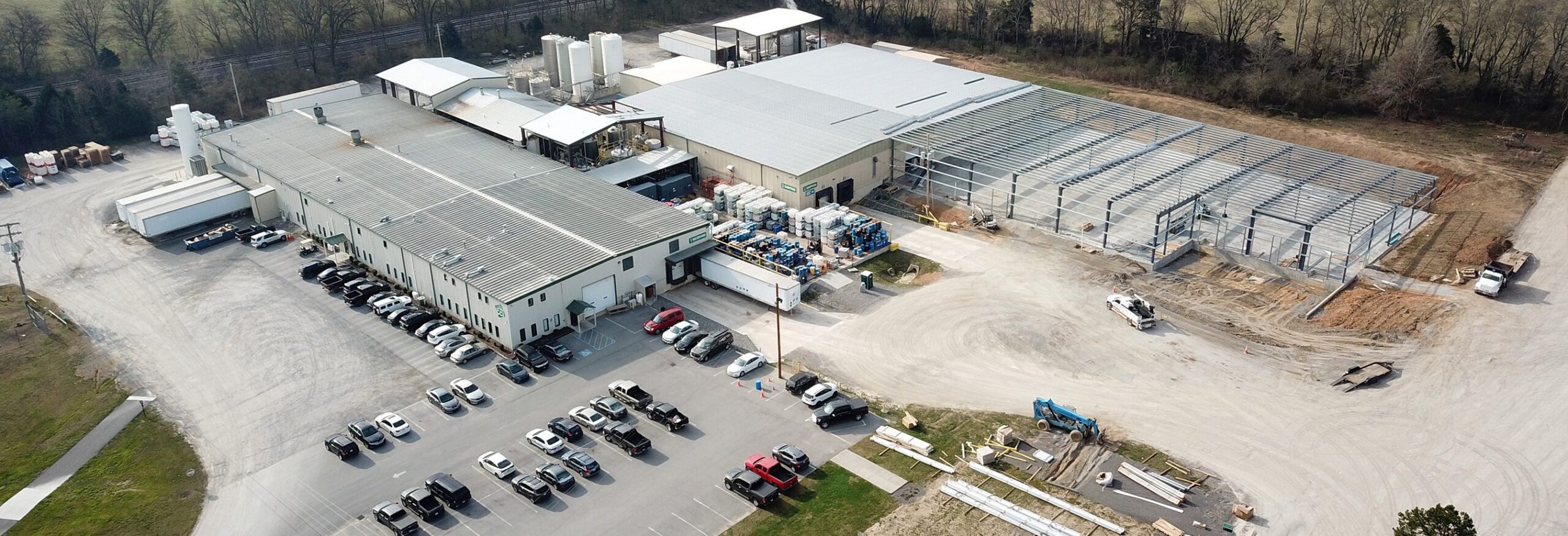 Aerial view of an industrial facility with multiple large buildings, parked cars, trucks, storage tanks, and a construction area with steel framework being erected next to a wooded area.