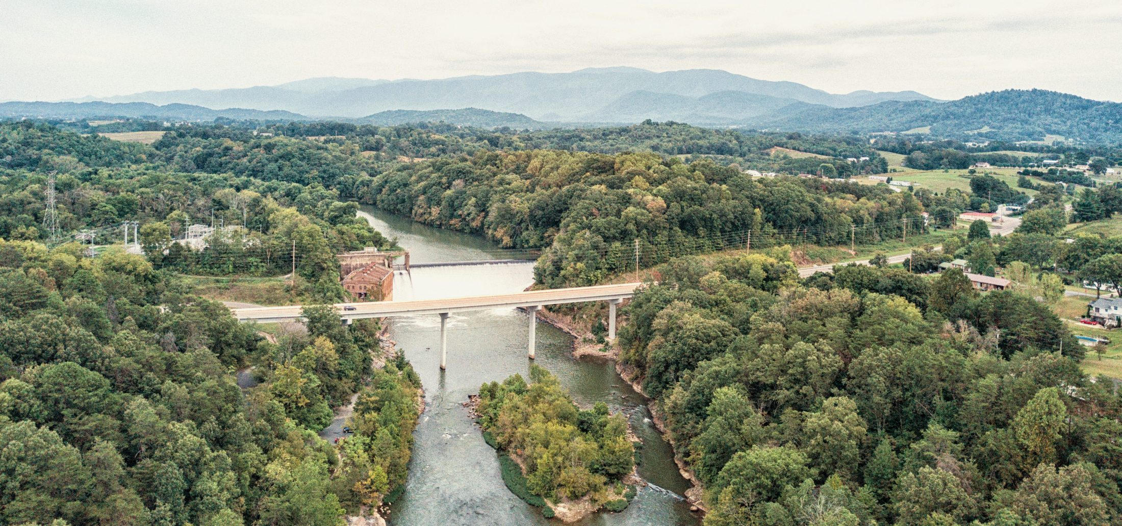 A wide aerial view of a bridge spanning a river, surrounded by dense green trees and rolling hills, with distant mountains under a cloudy sky.
