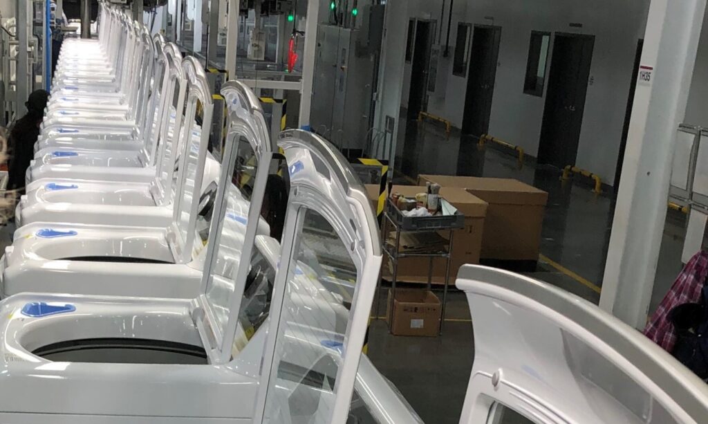 A row of white washing machines with open lids is lined up inside a factory or warehouse, with boxes and industrial equipment visible in the background.