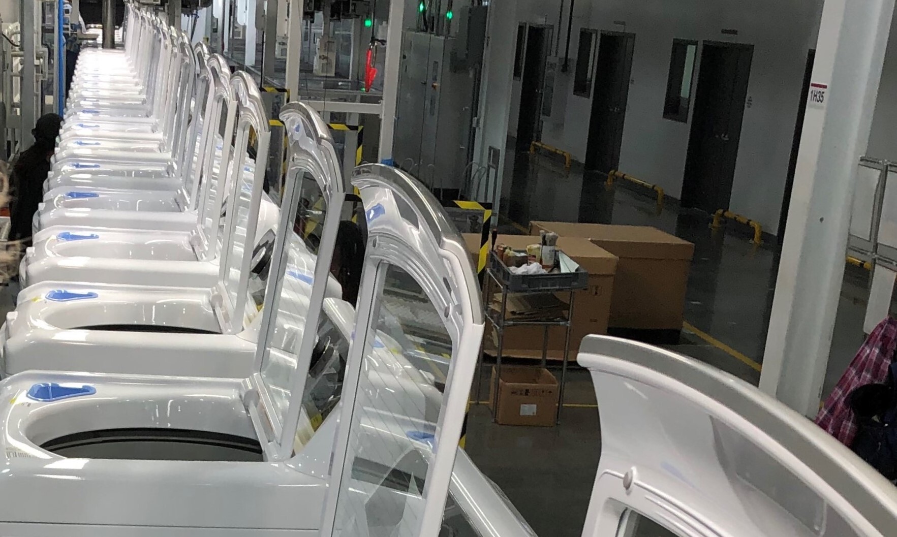A row of white washing machines with open lids is lined up inside a factory or warehouse, with boxes and industrial equipment visible in the background.