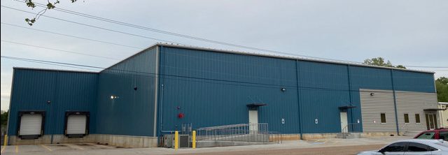 A large blue industrial warehouse with two loading docks, a ramp, and several doors. The building sits on a concrete lot with a few cars parked in front and trees visible in the background.