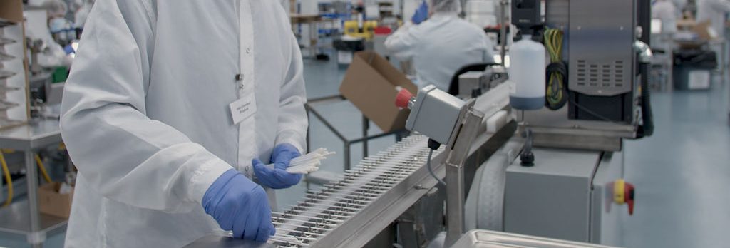 A person wearing a white lab coat, gloves, and hair covering works at an assembly line in a clean, sterile manufacturing facility, handling items arranged in rows.