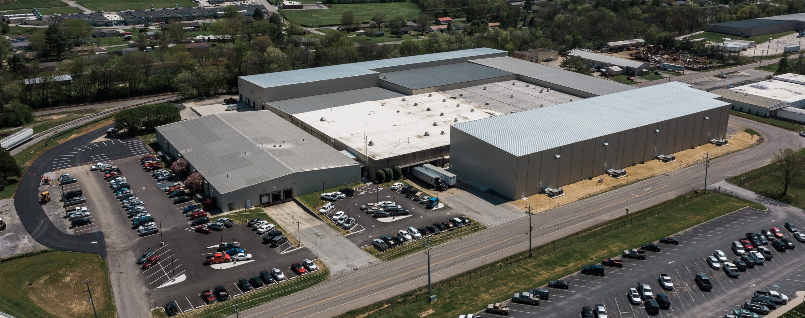 Aerial view of a large industrial warehouse complex with a white roof, surrounded by parking lots filled with cars, and green fields and roads nearby.
