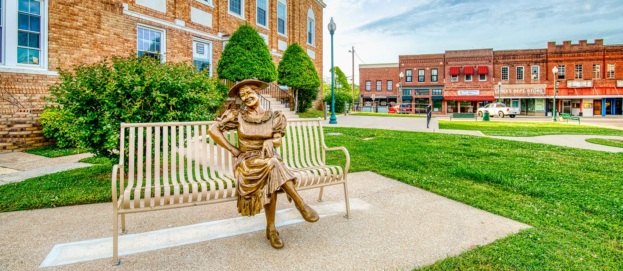 A bronze statue of a woman in a hat sits on a bench outside a brick building, with grassy lawns and historic storefronts visible across the street in a small town square.