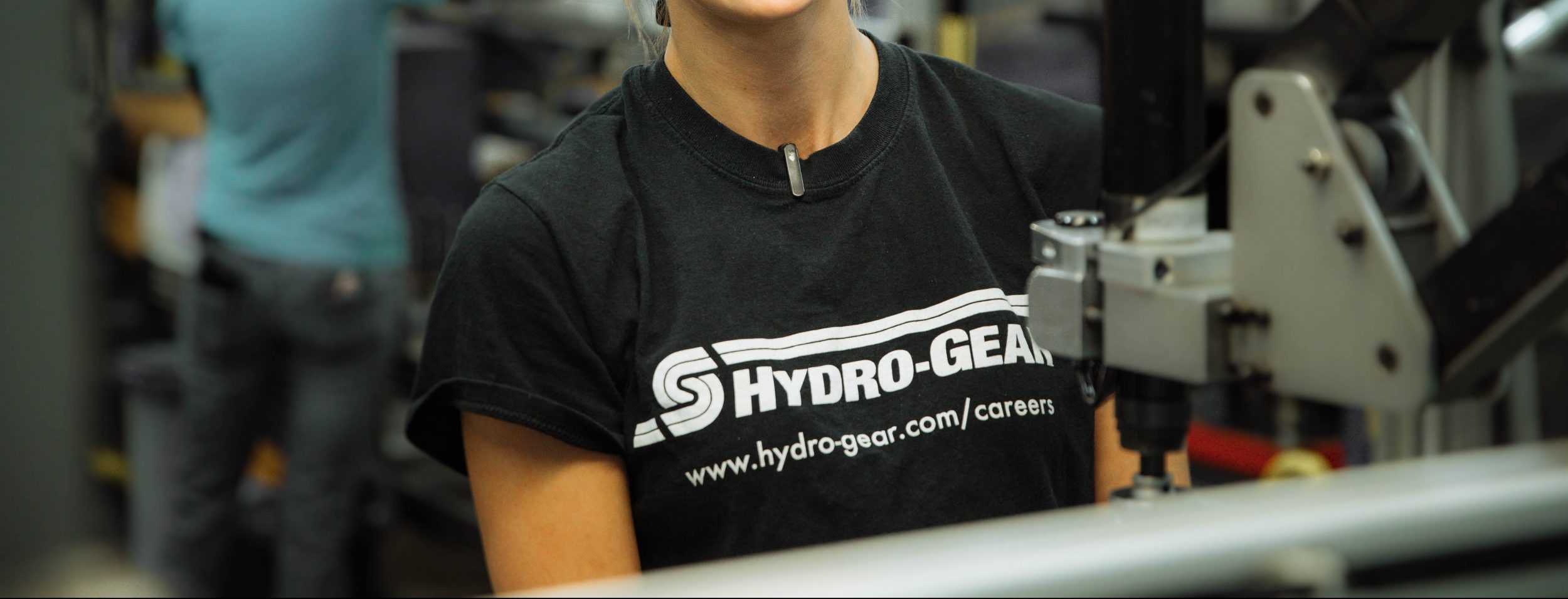 Person wearing a black Hydro-Gear t-shirt standing near machinery in what appears to be a workshop or industrial setting; only the upper torso and part of the face are visible.