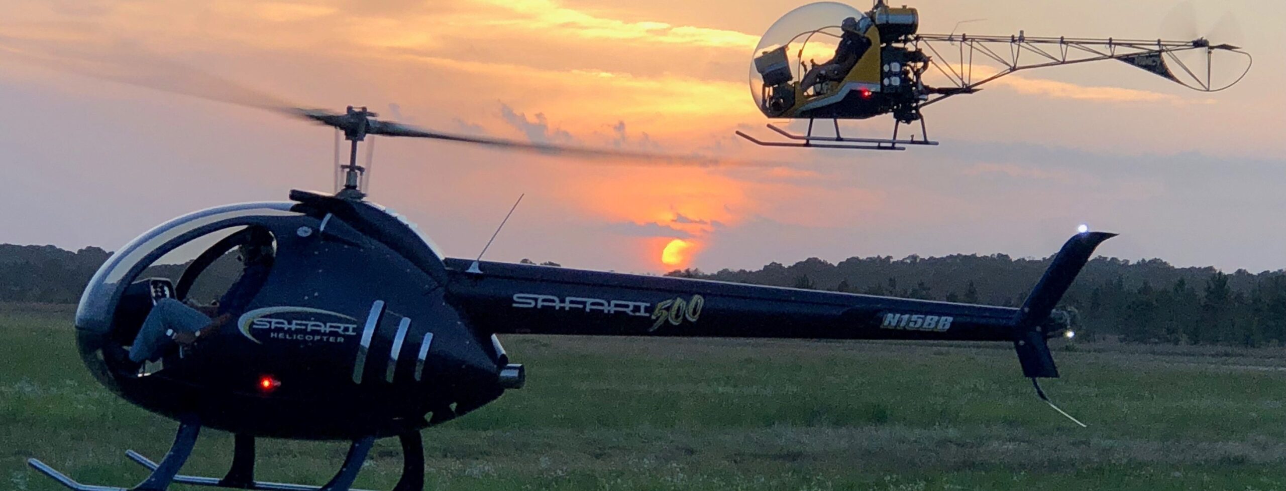 Two helicopters fly over a grassy field at sunset. The closer black helicopter is labeled “Safari 500,” while a smaller yellow helicopter flies in the background with the orange sun low on the horizon.