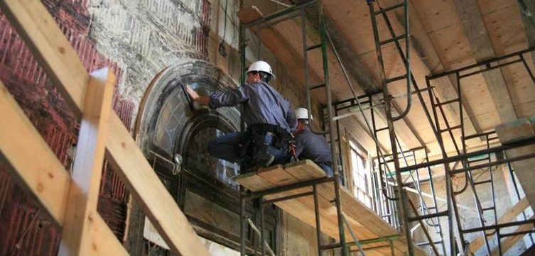 Two construction workers wearing hard hats and safety gear restore an ornate, arched architectural feature while standing on scaffolding inside a building under renovation.