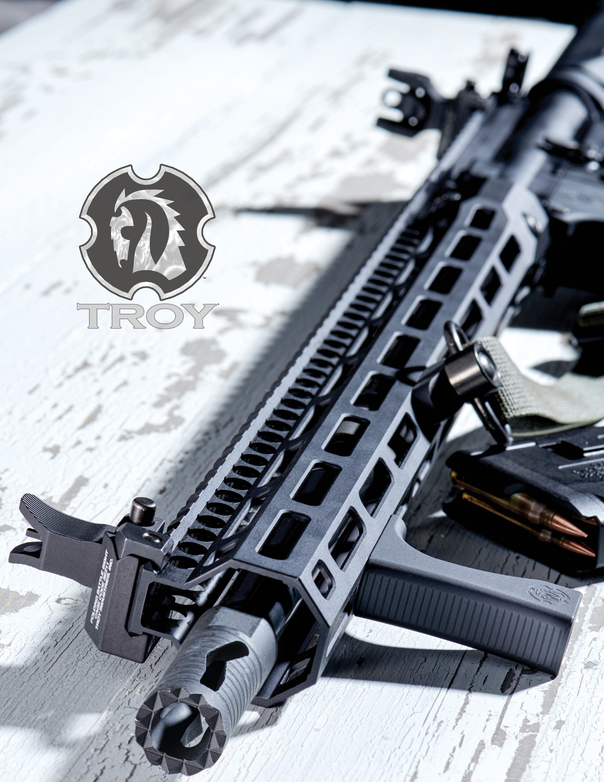 Close-up of a black tactical rifle with a textured handguard and Troy logo above it, placed on a distressed white wooden surface. Various firearm details and components are visible.