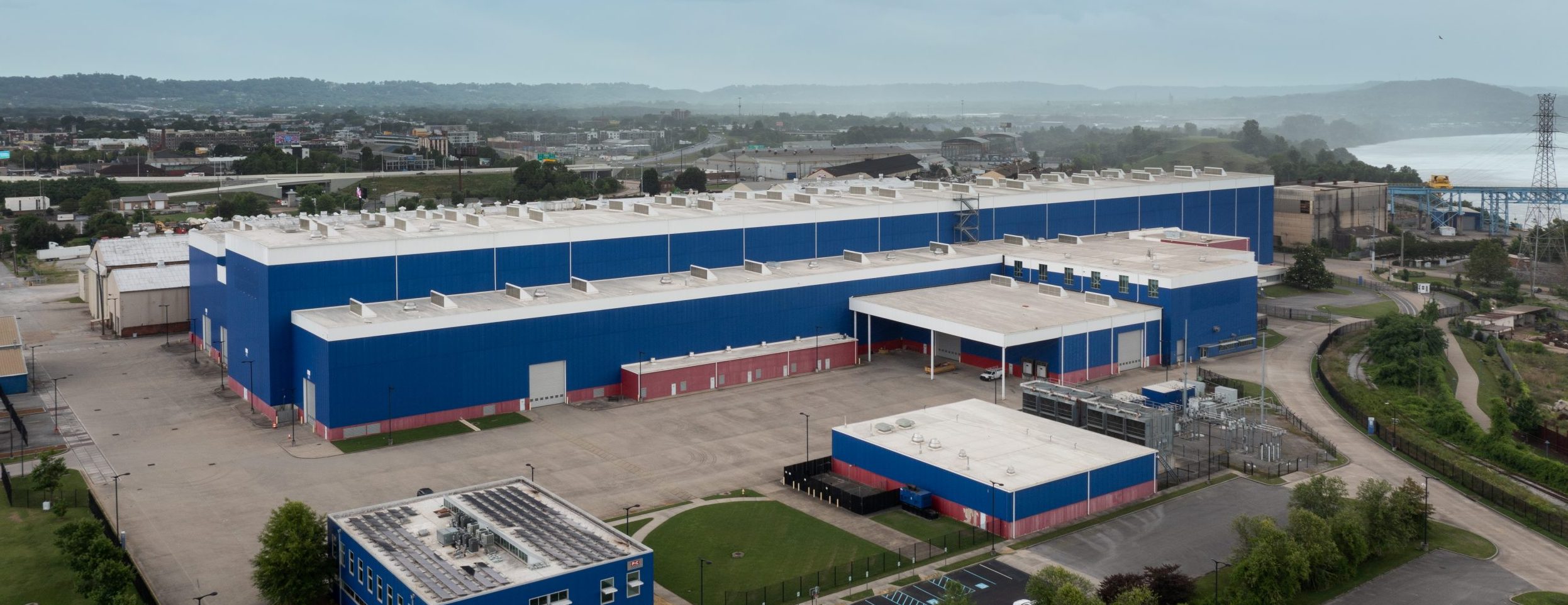 A large blue and red industrial building with several rooftop units sits in a spacious, paved area surrounded by greenery and roads, with a cityscape and hills visible in the background under a cloudy sky.