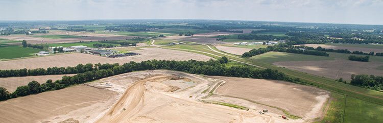 Aerial view of a large construction site surrounded by open fields and farmland, with roads, trees, and scattered buildings visible in the distance under a partly cloudy sky.