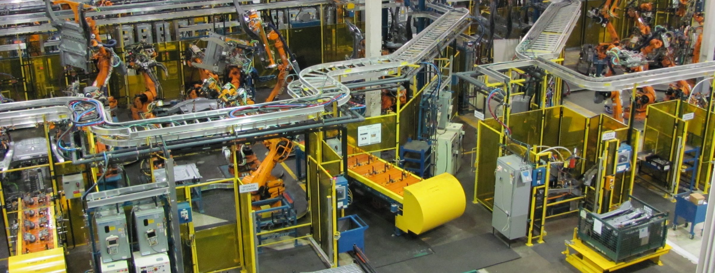 A busy factory floor with multiple orange robotic arms working on assembling machinery, surrounded by yellow safety barriers, conveyor belts, electrical panels, and various industrial equipment.