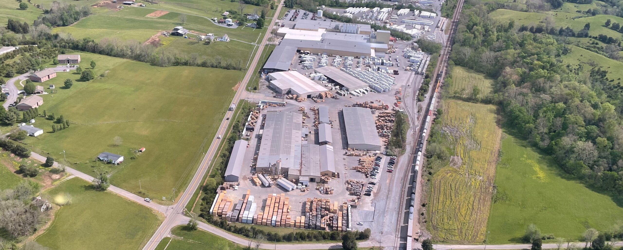 Aerial view of a large industrial lumber facility surrounded by green fields and scattered houses, with stacks of timber and buildings visible inside the complex. Roads and railroad tracks run alongside the site.