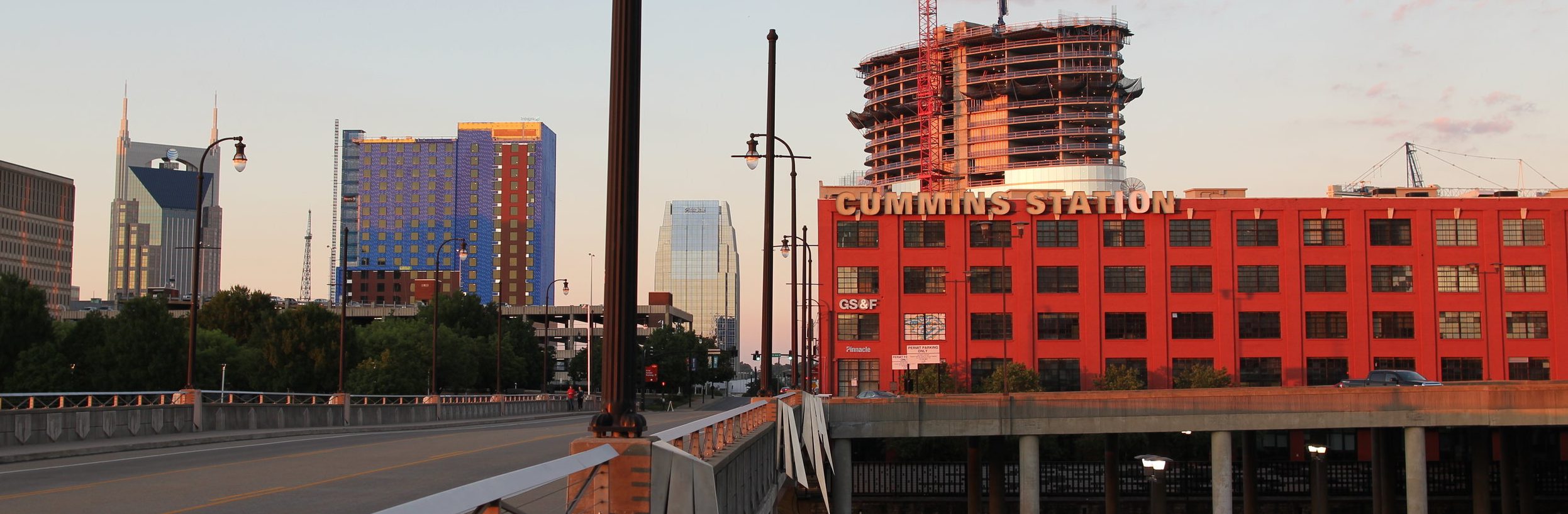 A cityscape at sunset with a red-brick building labeled "CUMMINS STATION," modern skyscrapers, a round building under construction, and a mostly empty road in the foreground.