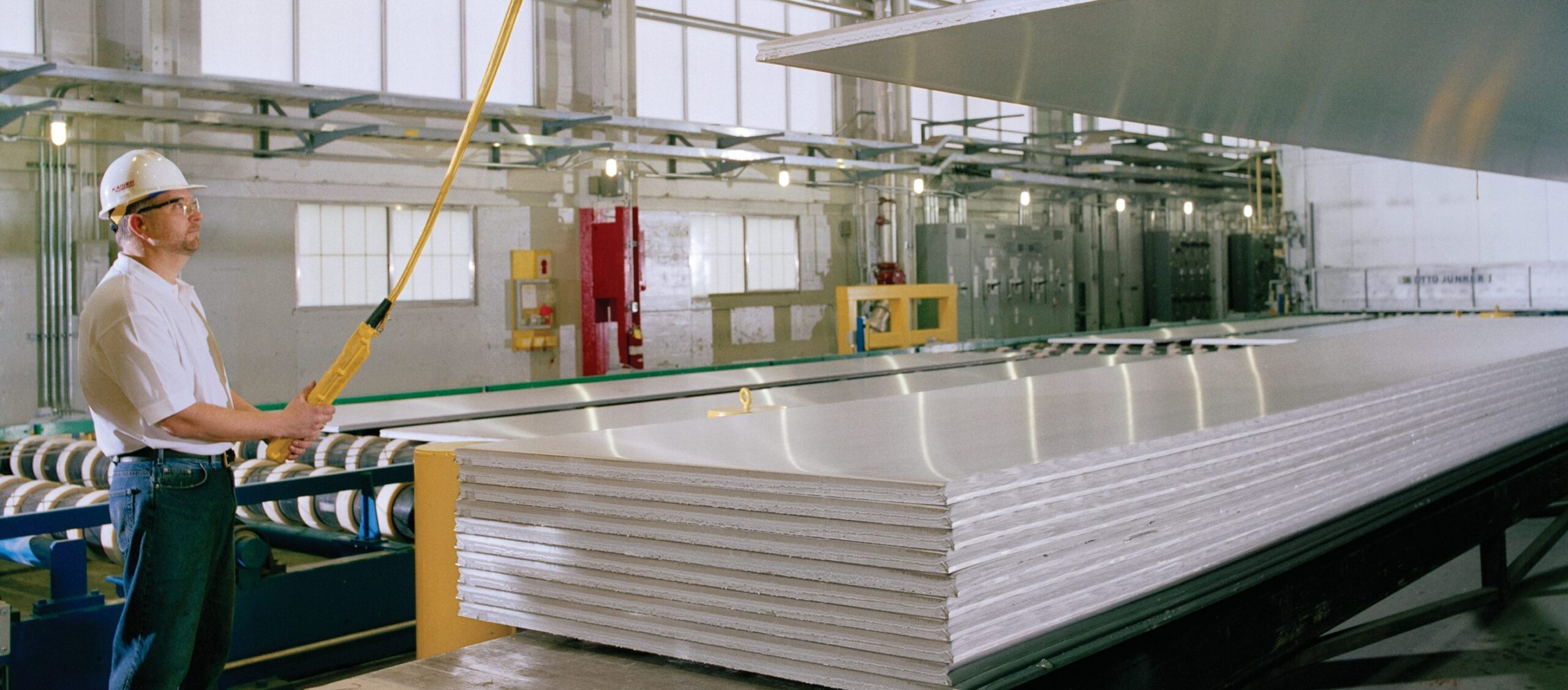 A worker in a hard hat and safety glasses operates machinery to lift a large metal sheet above a stack of similar sheets in an industrial factory setting.