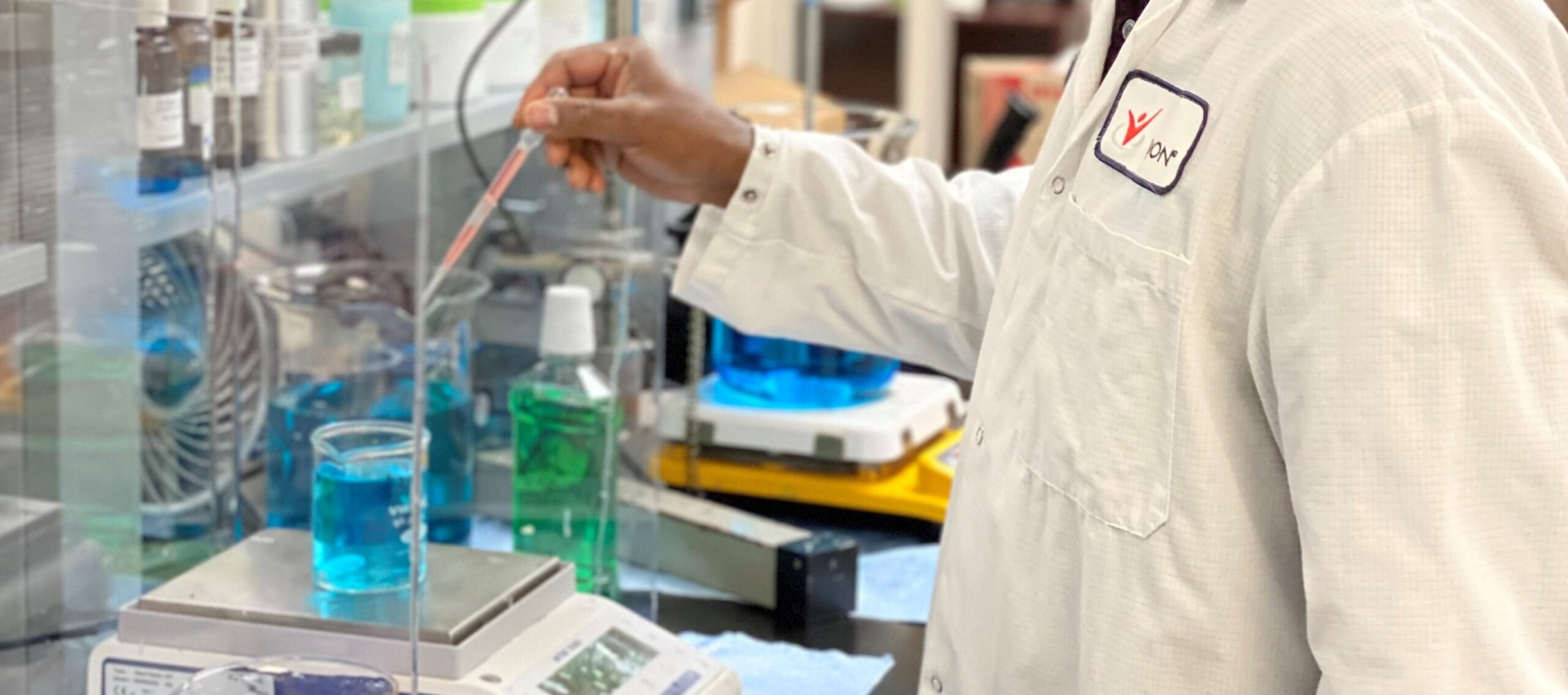 A person in a white lab coat uses a pipette to transfer liquid into a beaker containing blue solution on a digital scale in a laboratory setting. Shelves with lab equipment are visible in the background.