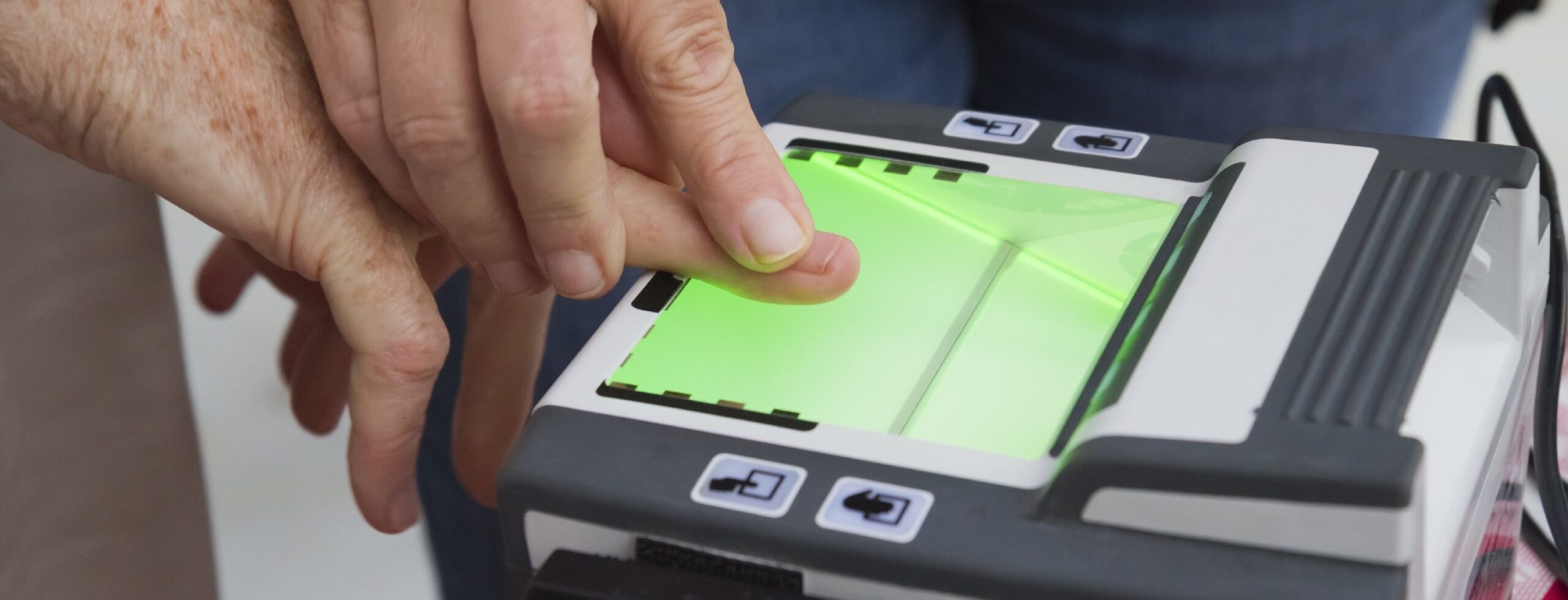 A close-up of a person's fingers being placed on a fingerprint scanner by another person, with the scanner surface glowing green.