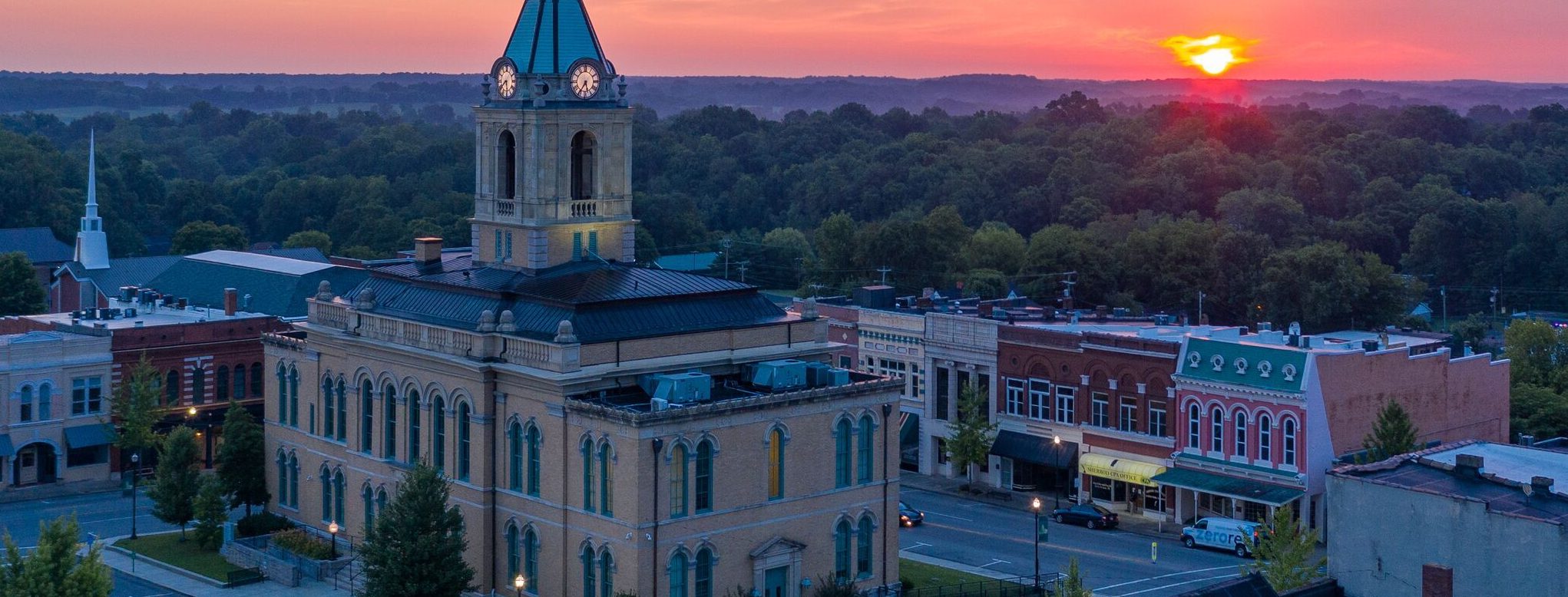 A historic clock tower building stands in a small downtown area at sunset, with the sun setting behind distant hills and trees, and colorful historic storefronts along the street.