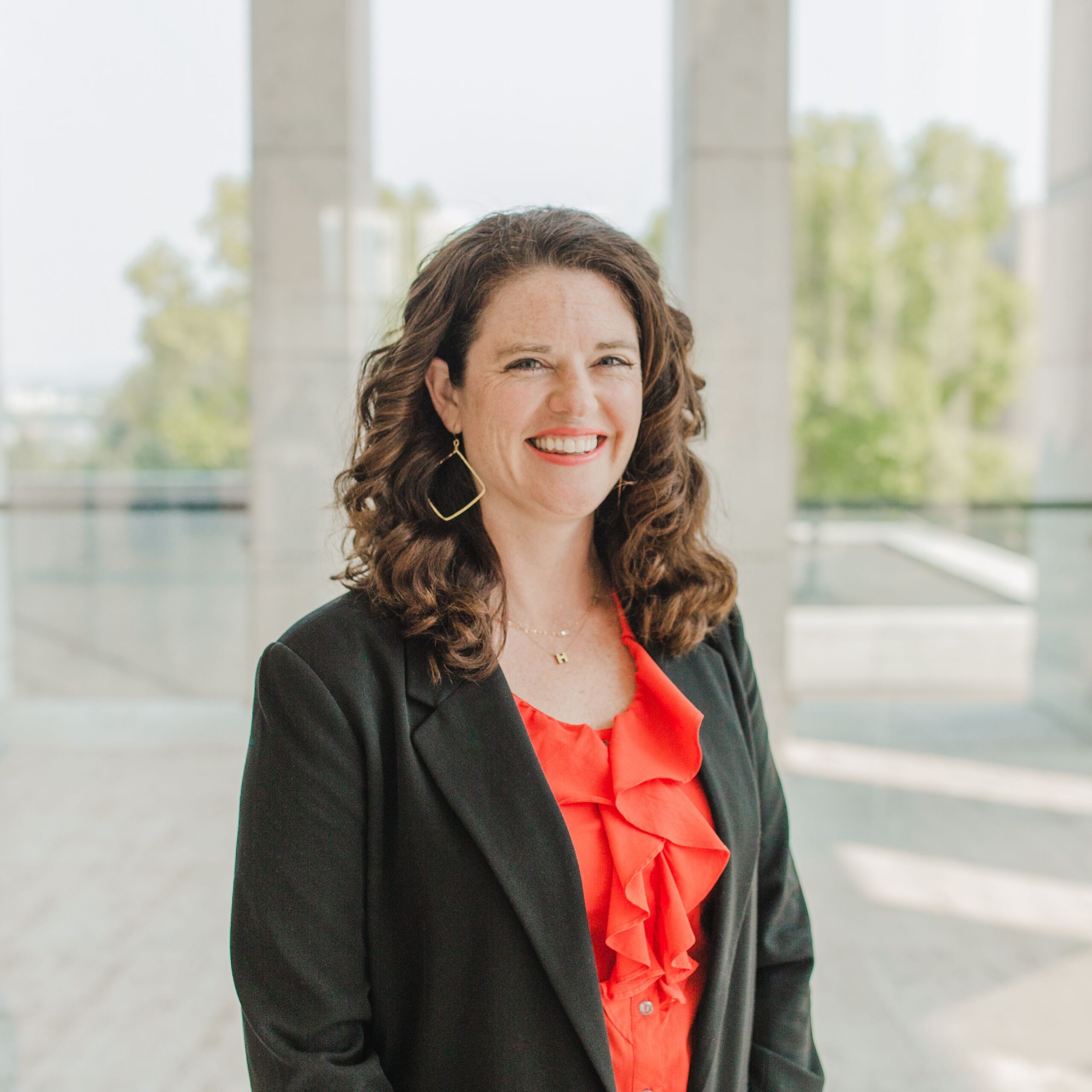 A woman with wavy brown hair, wearing a black blazer and a bright red ruffled blouse, stands smiling in a modern, sunlit building with large windows and distant greenery outside.