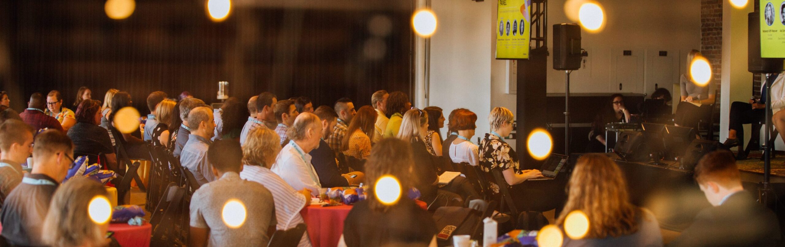 A group of people seated at round tables attentively listens to a speaker at a conference or seminar, with warm string lights blurred in the foreground and presentation slides visible in the background.