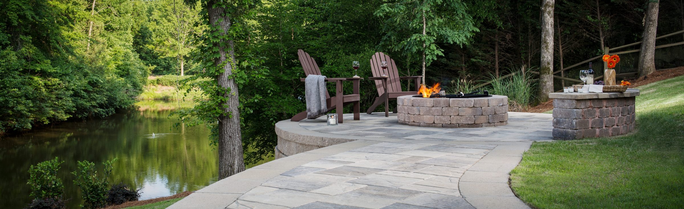A stone patio with Adirondack chairs surrounds a fire pit next to a peaceful pond, bordered by lush green trees and grass, creating a relaxing outdoor setting.