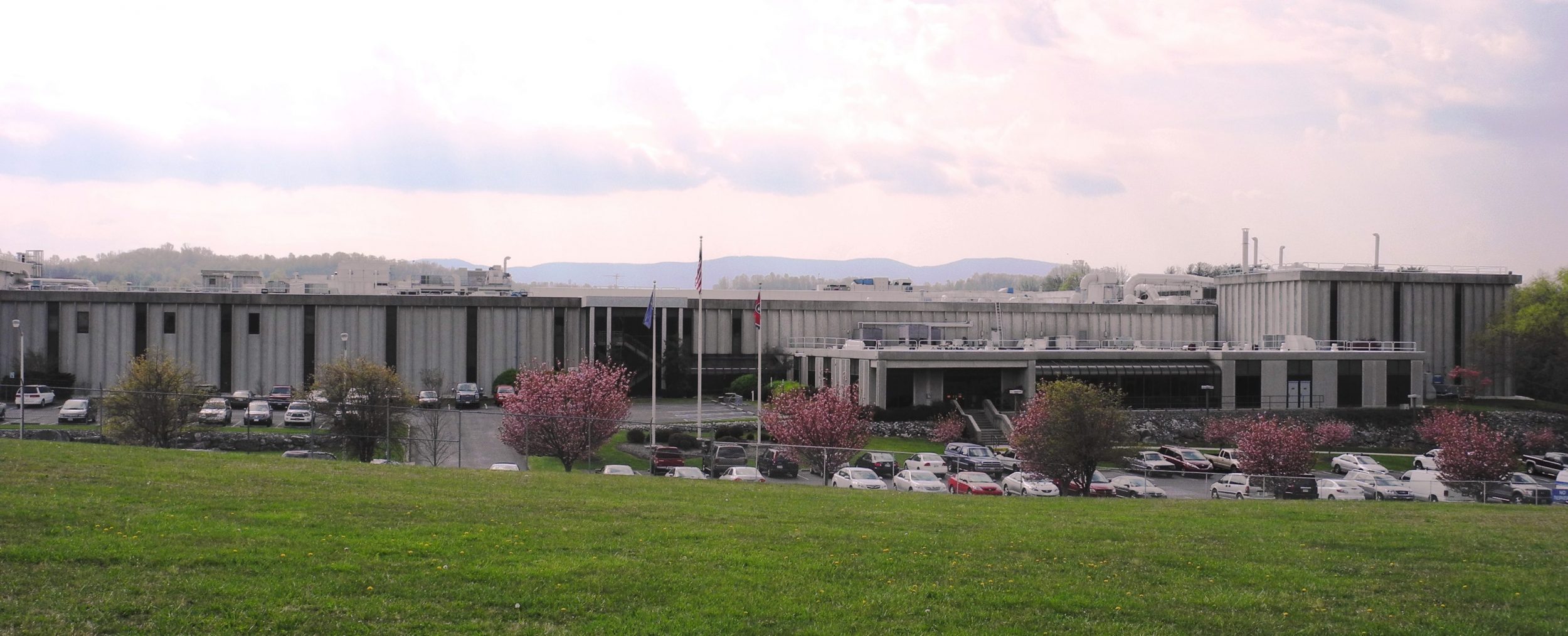 A large, low-rise building with a flat roof sits behind a parking lot filled with cars and surrounded by small blooming trees; mountains and a cloudy sky are visible in the background.